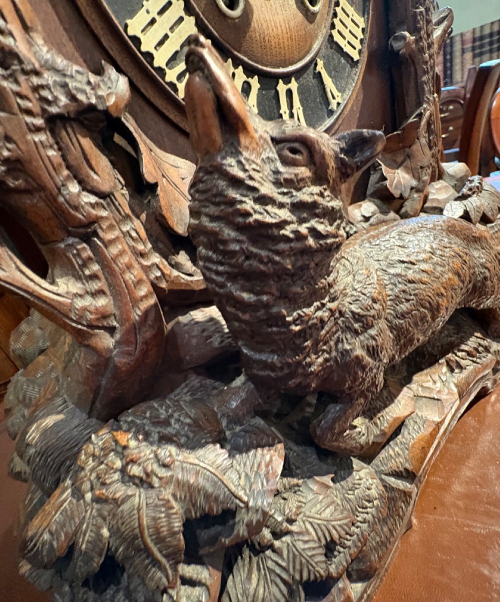 An intricately hand-carved dark wood Black Forest cuckoo clock featuring a bird atop a nest, a framed dial with Roman numerals, and a fox figure at the base. View XI