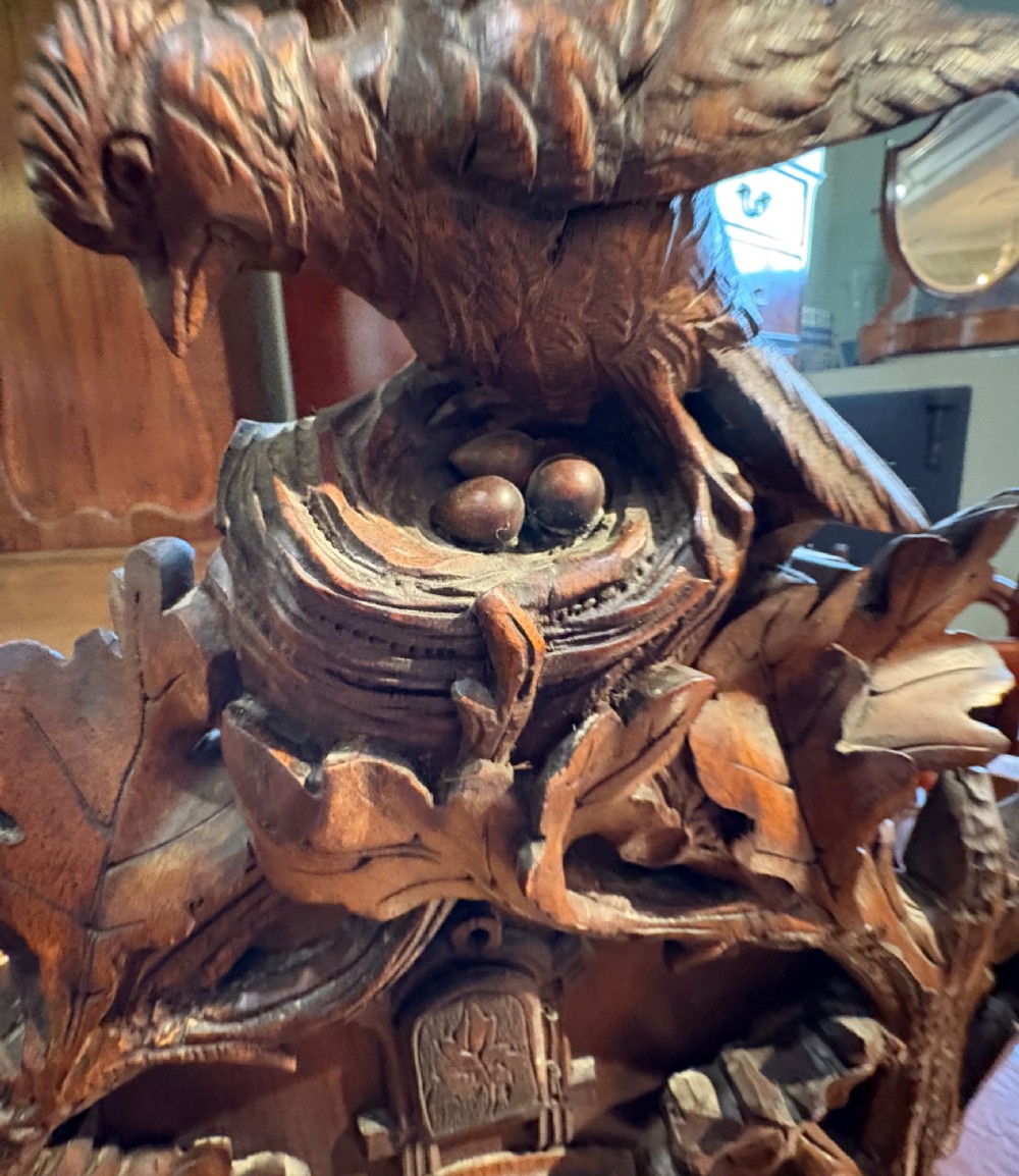 An intricately hand-carved dark wood Black Forest cuckoo clock featuring a bird atop a nest, a framed dial with Roman numerals, and a fox figure at the base. View XII
