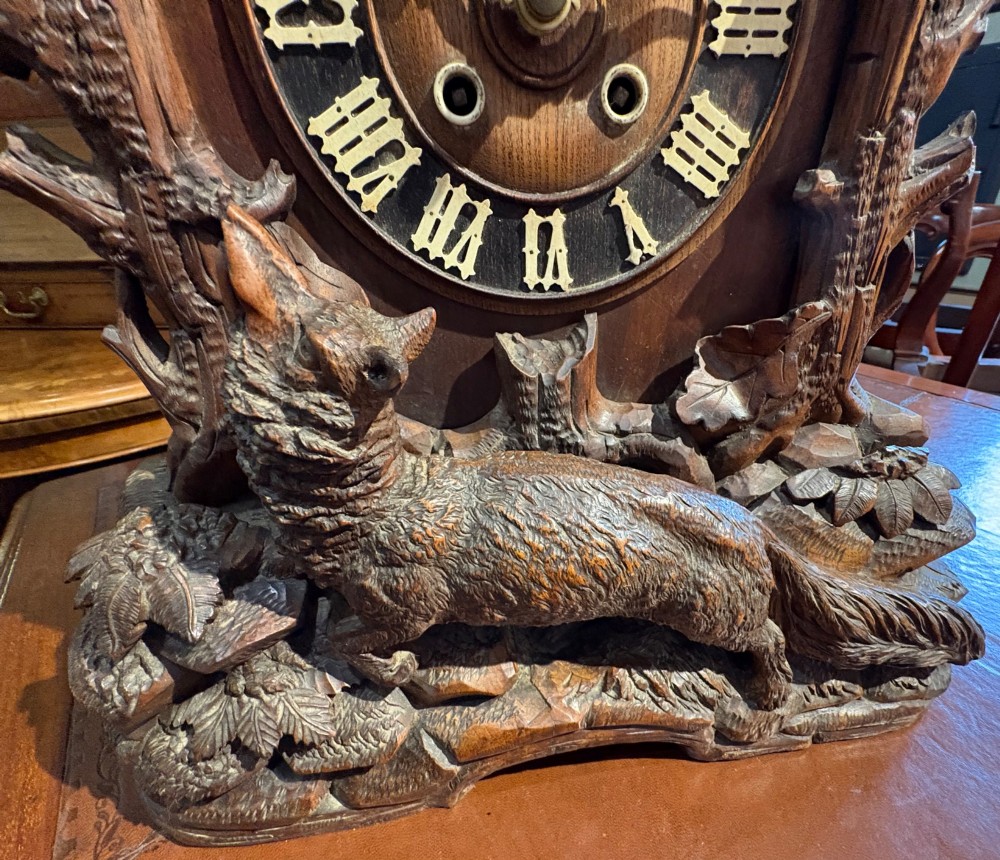 An intricately hand-carved dark wood Black Forest cuckoo clock featuring a bird atop a nest, a framed dial with Roman numerals, and a fox figure at the base. View XIII