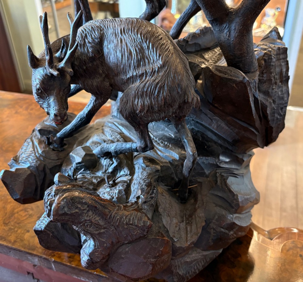 A dark hand-carved wooden Black Forest sculpture depicting two horned animals positioned on a craggy rocky outcrop beside a gnarled tree branch. View II