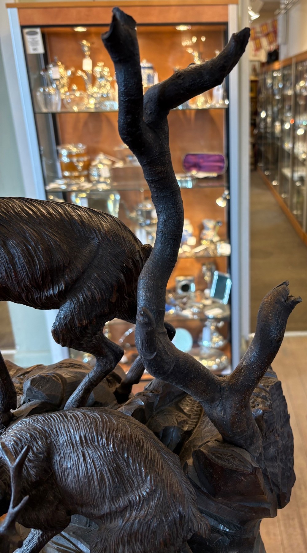 A dark hand-carved wooden Black Forest sculpture depicting two horned animals positioned on a craggy rocky outcrop beside a gnarled tree branch. View V