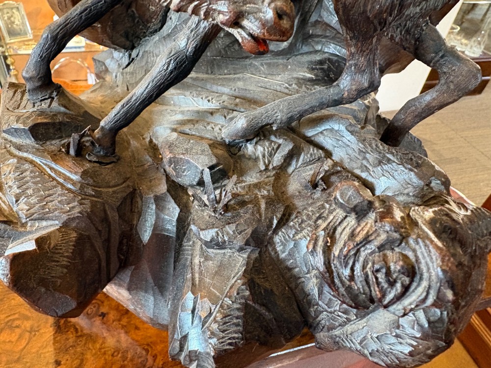 A dark hand-carved wooden Black Forest sculpture depicting two horned animals positioned on a craggy rocky outcrop beside a gnarled tree branch. View VI