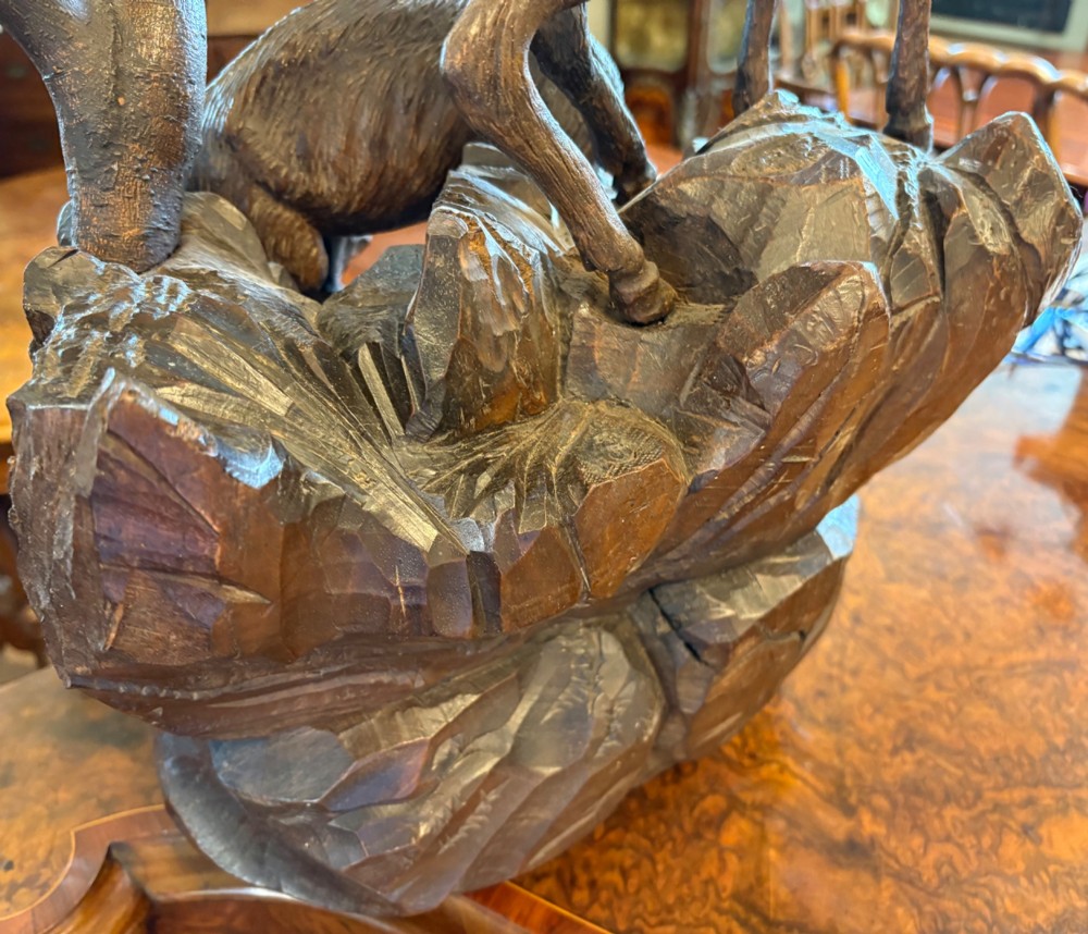 A dark hand-carved wooden Black Forest sculpture depicting two horned animals positioned on a craggy rocky outcrop beside a gnarled tree branch. View VIII