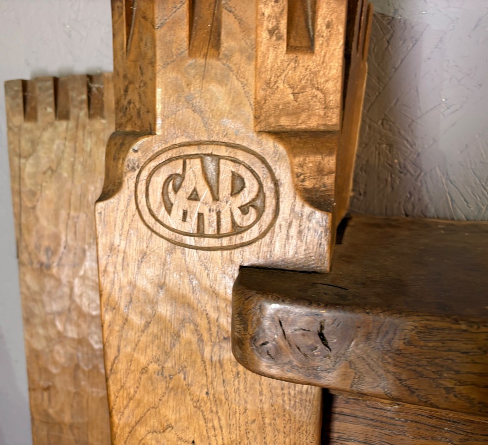A hand-carved solid oak fire surround featuring an adzed finish, castellated top posts, carved monograms, and small side shelves. View IV