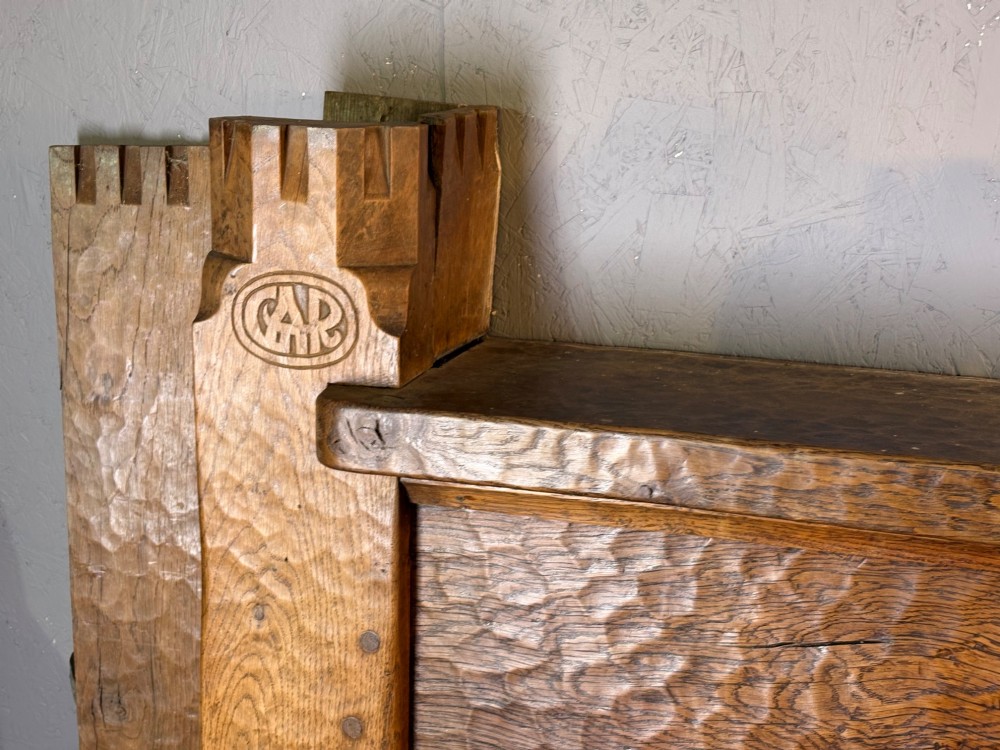 A hand-carved solid oak fire surround featuring an adzed finish, castellated top posts, carved monograms, and small side shelves. View V