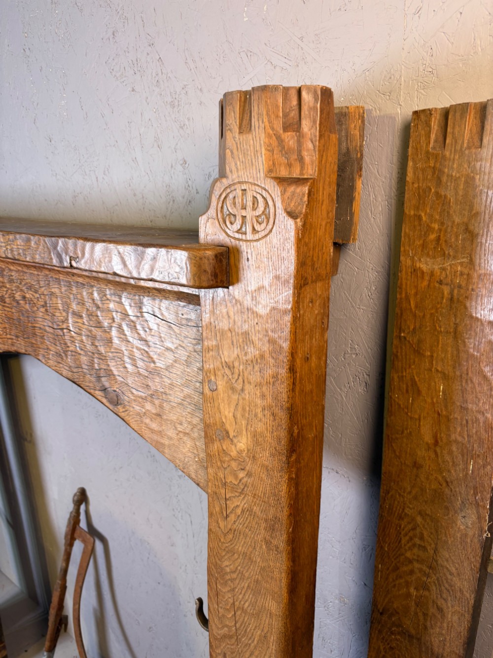 A hand-carved solid oak fire surround featuring an adzed finish, castellated top posts, carved monograms, and small side shelves. View XIV