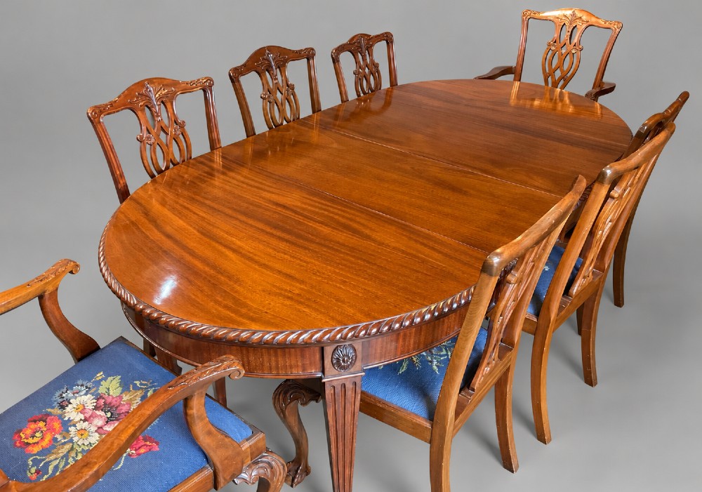 An oval mahogany extending dining table with a carved edge and winding mechanism, surrounded by eight carved wooden chairs featuring blue floral needlepoint seats.