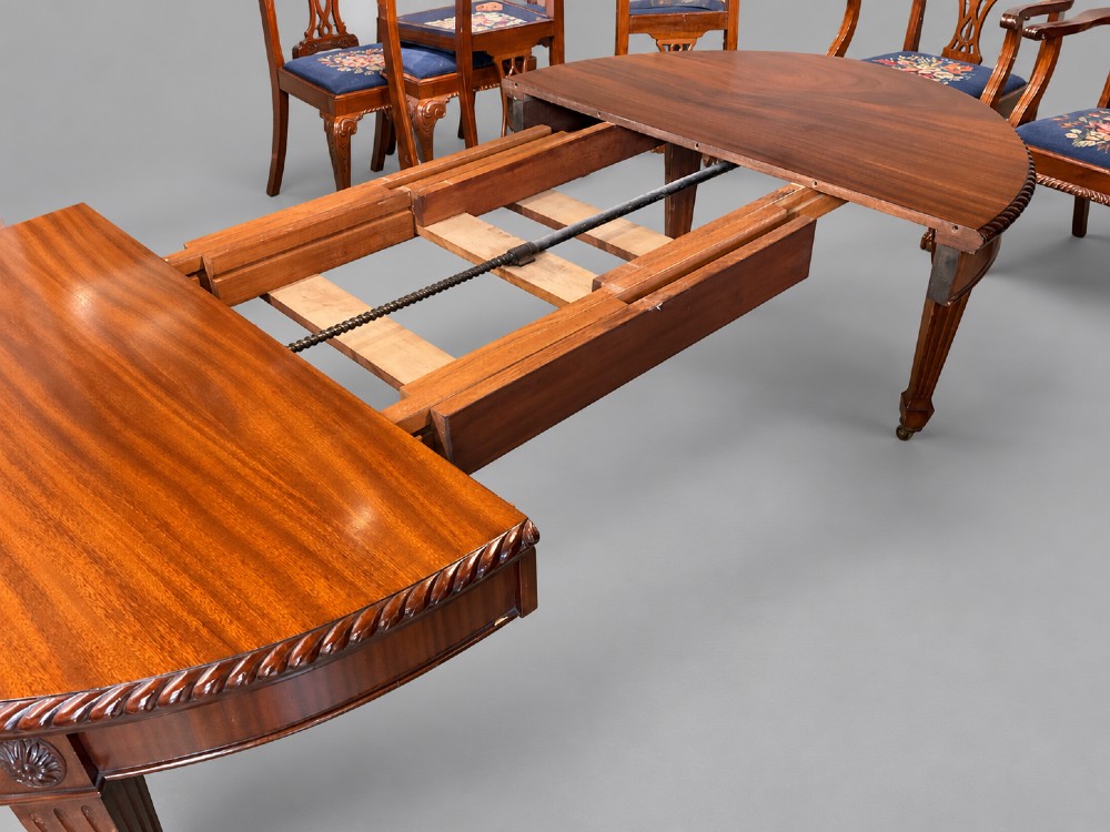 An oval mahogany extending dining table with a carved edge and winding mechanism, surrounded by eight carved wooden chairs featuring blue floral needlepoint seats. View III