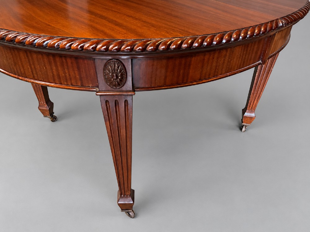 An oval mahogany extending dining table with a carved edge and winding mechanism, surrounded by eight carved wooden chairs featuring blue floral needlepoint seats. View IV
