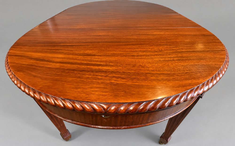 An oval mahogany extending dining table with a carved edge and winding mechanism, surrounded by eight carved wooden chairs featuring blue floral needlepoint seats. View VI