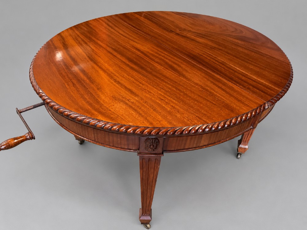 An oval mahogany extending dining table with a carved edge and winding mechanism, surrounded by eight carved wooden chairs featuring blue floral needlepoint seats. View VII