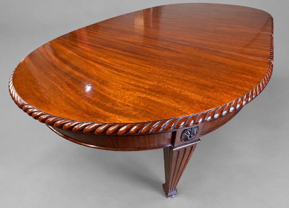 An oval mahogany extending dining table with a carved edge and winding mechanism, surrounded by eight carved wooden chairs featuring blue floral needlepoint seats. View VIII