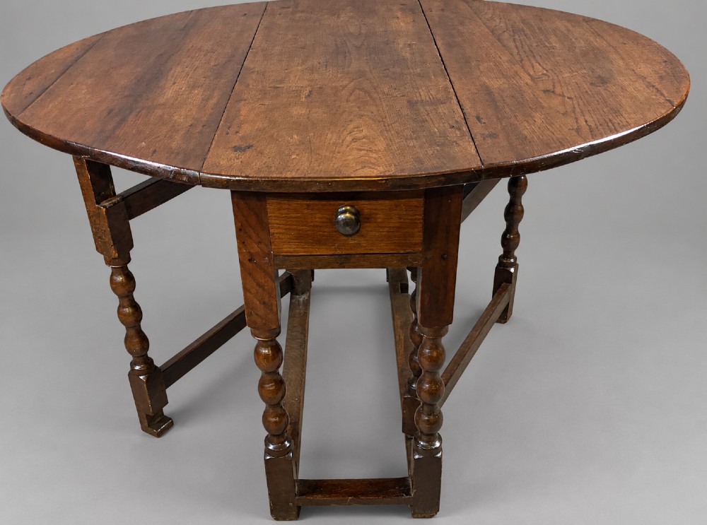 A 17th-century oak gateleg dining table with an oval drop-leaf top, turned legs, a box stretcher base, and a small end drawer.