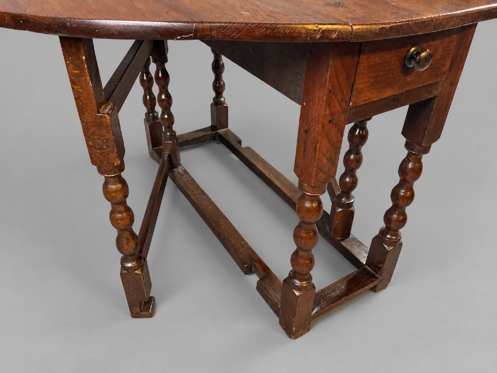 A 17th-century oak gateleg dining table with an oval drop-leaf top, turned legs, a box stretcher base, and a small end drawer. View IV