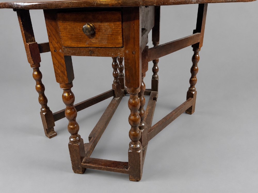 A 17th-century oak gateleg dining table with an oval drop-leaf top, turned legs, a box stretcher base, and a small end drawer. View V