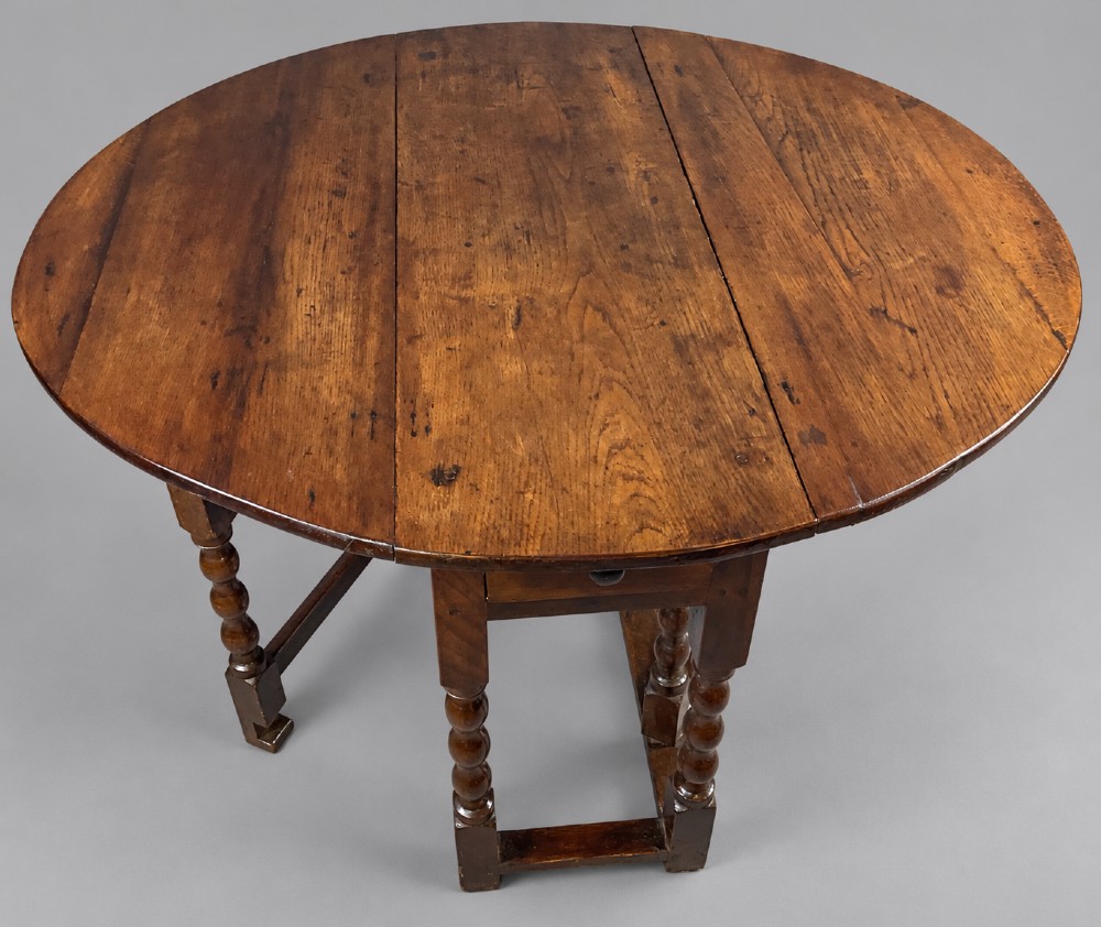 A 17th-century oak gateleg dining table with an oval drop-leaf top, turned legs, a box stretcher base, and a small end drawer. View VI