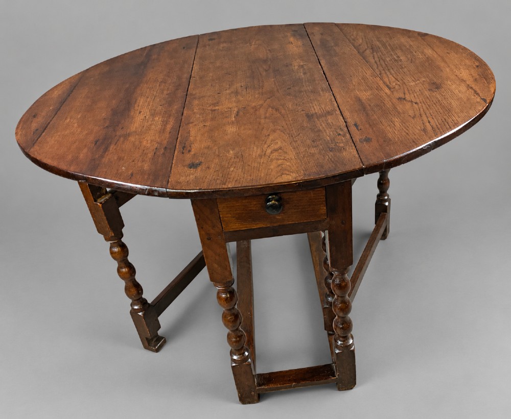 A 17th-century oak gateleg dining table with an oval drop-leaf top, turned legs, a box stretcher base, and a small end drawer. View VII