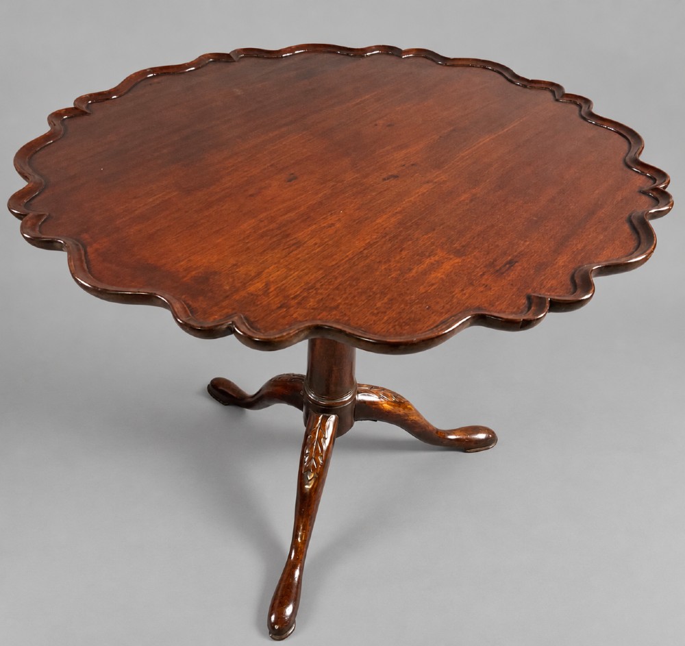 An 18th-century mahogany tilt-top supper table positioned vertically, displaying a scalloped piecrust edge rim and a turned tripod base with carved cabriole legs. View II