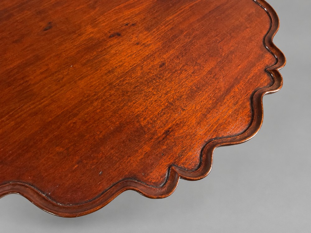 An 18th-century mahogany tilt-top supper table positioned vertically, displaying a scalloped piecrust edge rim and a turned tripod base with carved cabriole legs. View IV