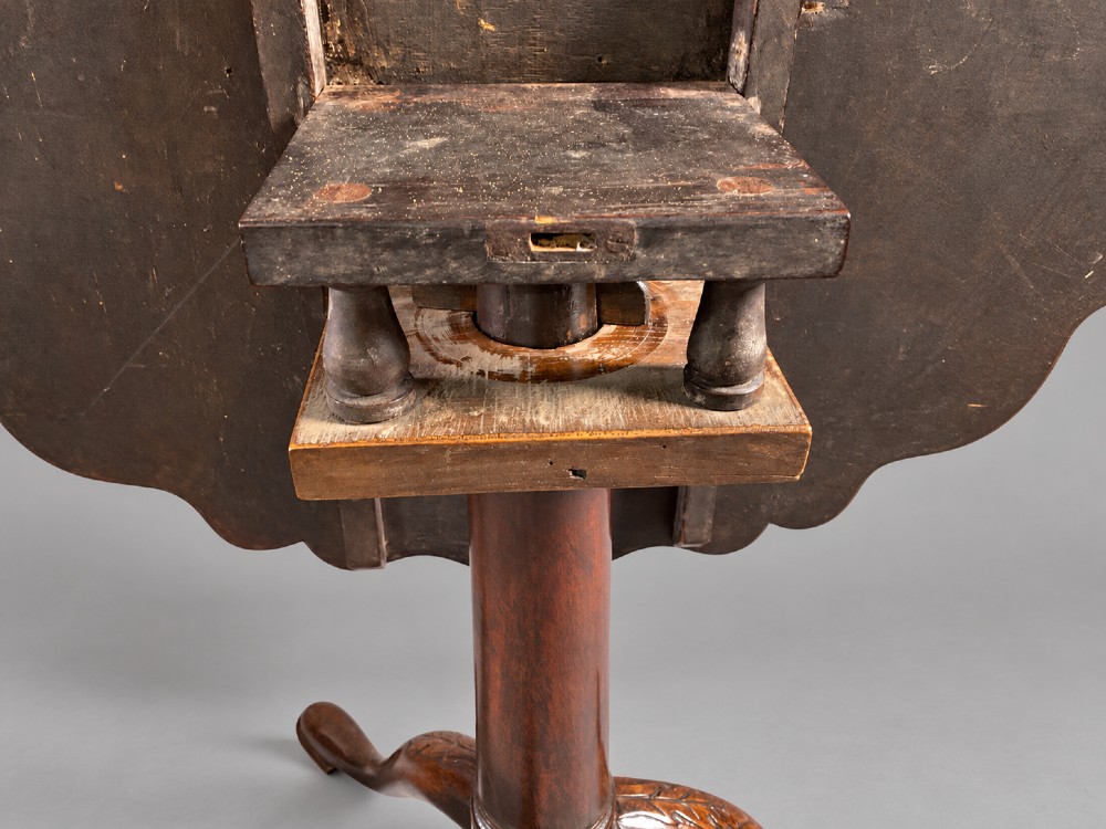 An 18th-century mahogany tilt-top supper table positioned vertically, displaying a scalloped piecrust edge rim and a turned tripod base with carved cabriole legs. View V