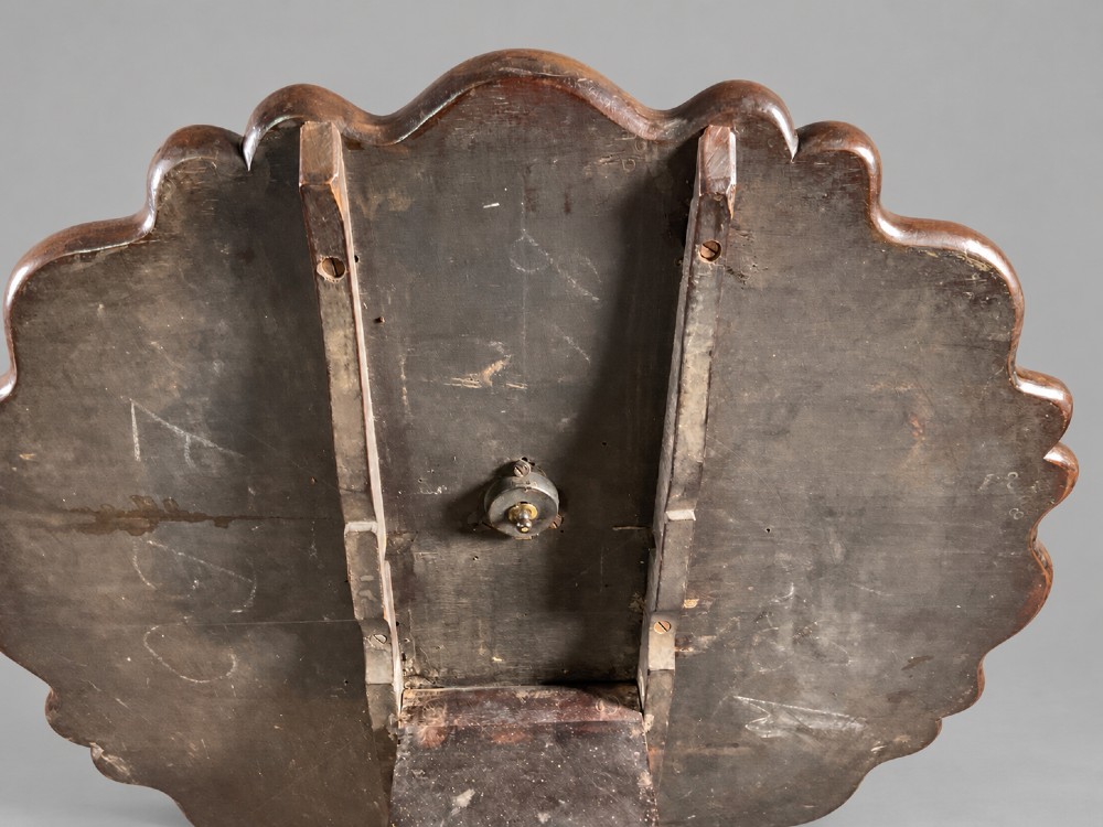 An 18th-century mahogany tilt-top supper table positioned vertically, displaying a scalloped piecrust edge rim and a turned tripod base with carved cabriole legs. View VI