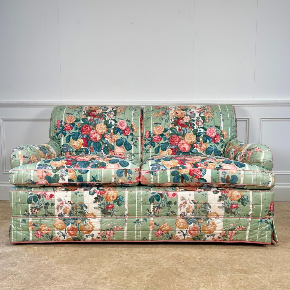 A two-seater upholstered sofa in sage green floral chintz fabric featuring a quilted texture and a fully skirted base.