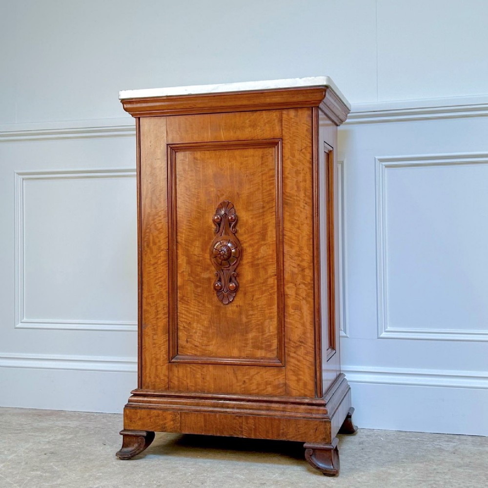 A Victorian satinwood pedestal cabinet with a white marble top and a single carved panelled door resting on a plinth base.