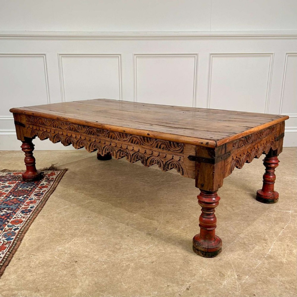 A low 19th-century Indian Takhat coffee table crafted from warm brown timber featuring a carved scalloped apron, iron corner brackets, and red turned legs.