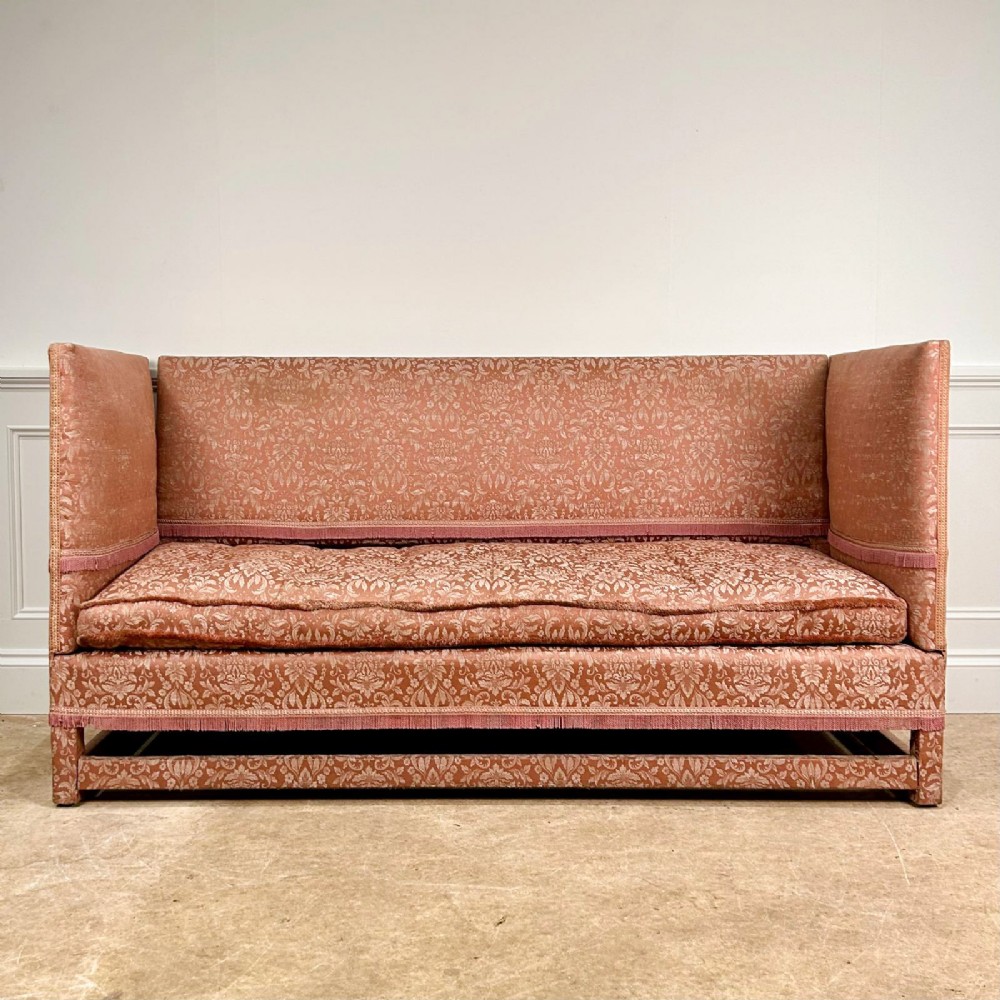 An Edwardian Knole sofa upholstered in rose pink damask fabric featuring high straight sides, a high back, and fringe trim along the borders.