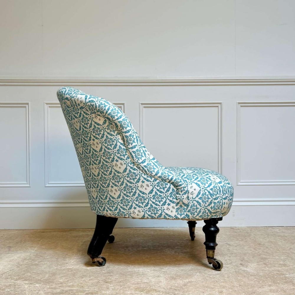 A low 19th-century armless slipper chair with blue and white botanical patterned upholstery, deep buttoned backrest, turned front legs, and brass castors. View II