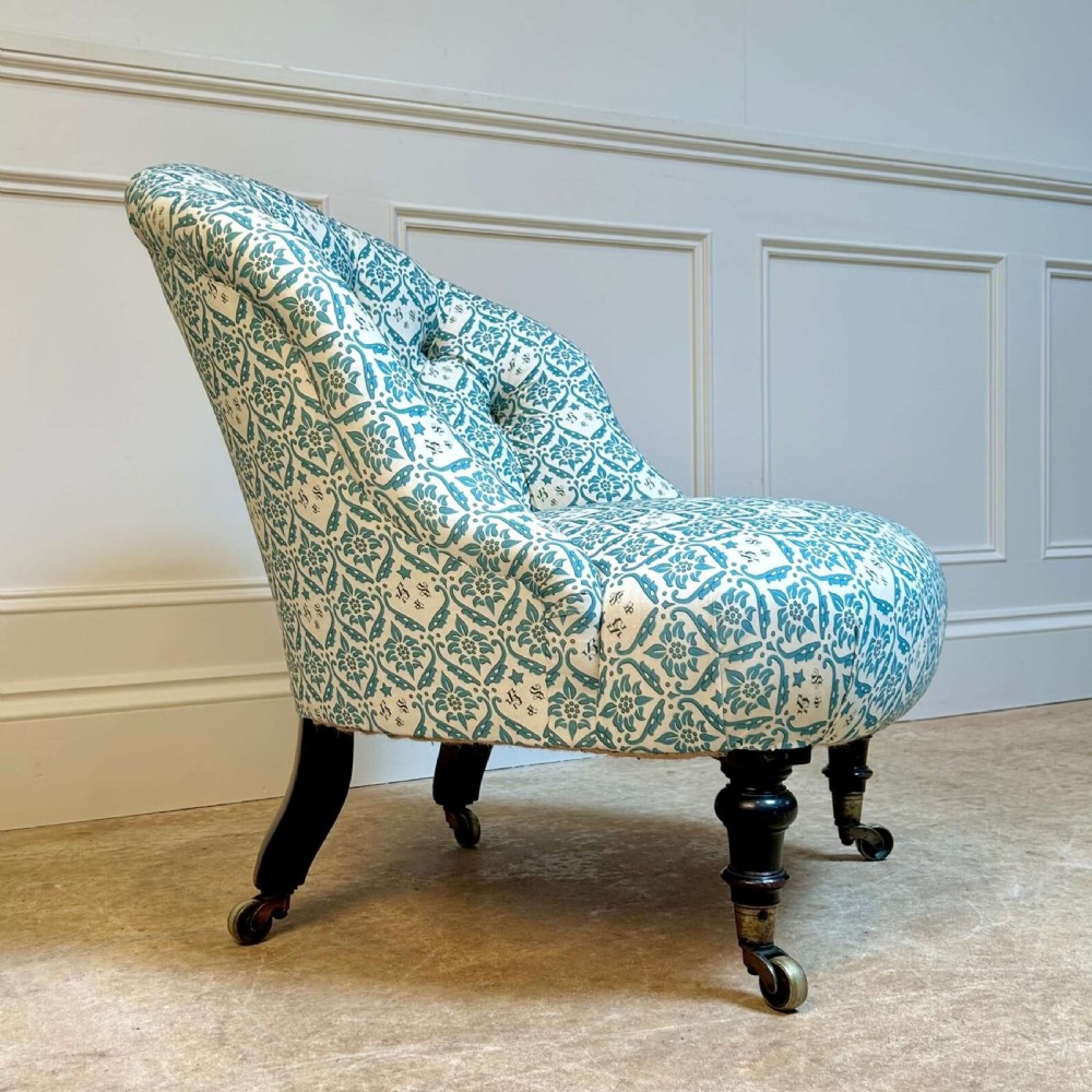 A low 19th-century armless slipper chair with blue and white botanical patterned upholstery, deep buttoned backrest, turned front legs, and brass castors. View IV