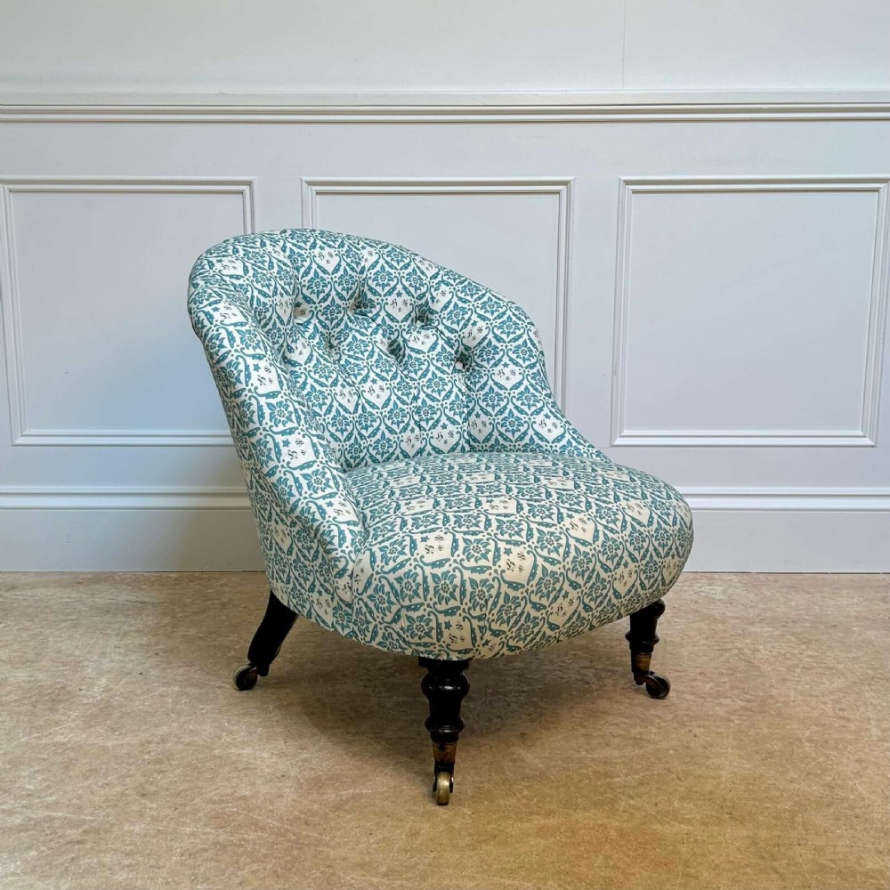 A low 19th-century armless slipper chair with blue and white botanical patterned upholstery, deep buttoned backrest, turned front legs, and brass castors. View V