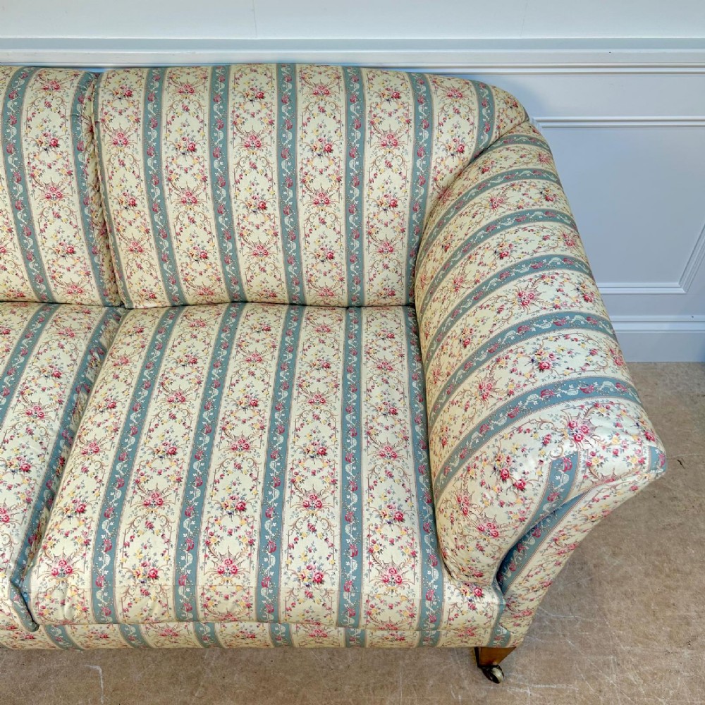 A two-seater Howard & Sons Chaplin sofa upholstered in cream, blue-striped, and floral patterned fabric with tapered wooden legs on brass castors. View V
