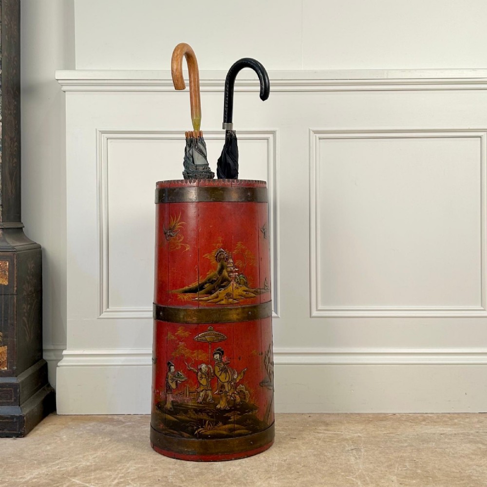A cylindrical red Edwardian umbrella stand featuring hand-painted gold Chinoiserie scenes and bound by brass hoops.