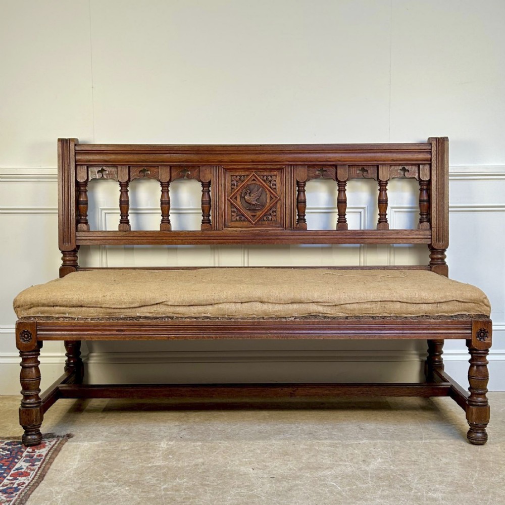 A 19th-century Gothic Revival oak hall bench featuring a backrest with turned spindles, carved arches, a central bird medallion, and a cushioned seat upholstered in natural hessian fabric.