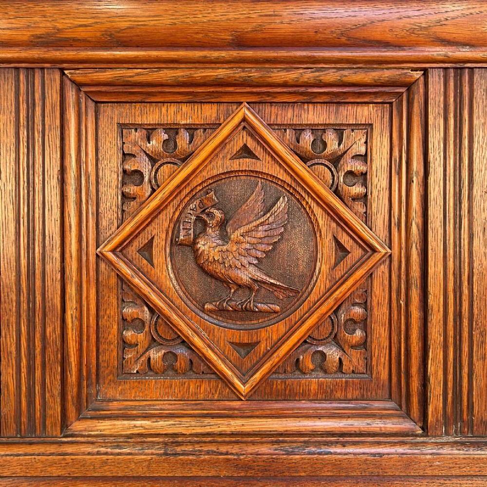 A 19th-century Gothic Revival oak hall bench featuring a backrest with turned spindles, carved arches, a central bird medallion, and a cushioned seat upholstered in natural hessian fabric. View II