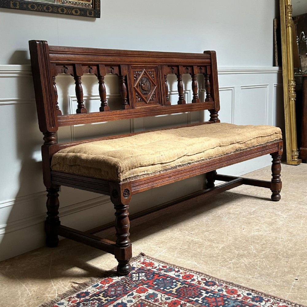 A 19th-century Gothic Revival oak hall bench featuring a backrest with turned spindles, carved arches, a central bird medallion, and a cushioned seat upholstered in natural hessian fabric. View III