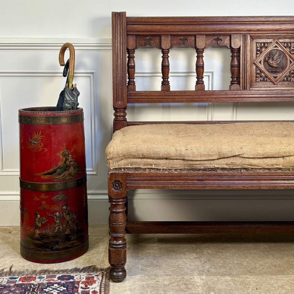 A 19th-century Gothic Revival oak hall bench featuring a backrest with turned spindles, carved arches, a central bird medallion, and a cushioned seat upholstered in natural hessian fabric. View IV