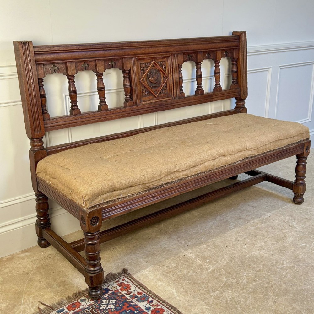A 19th-century Gothic Revival oak hall bench featuring a backrest with turned spindles, carved arches, a central bird medallion, and a cushioned seat upholstered in natural hessian fabric. View V