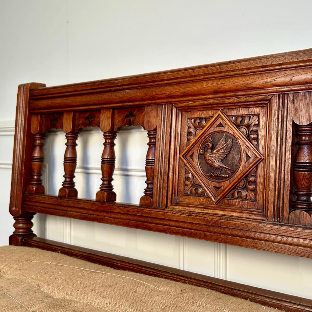 A 19th-century Gothic Revival oak hall bench featuring a backrest with turned spindles, carved arches, a central bird medallion, and a cushioned seat upholstered in natural hessian fabric. View VI