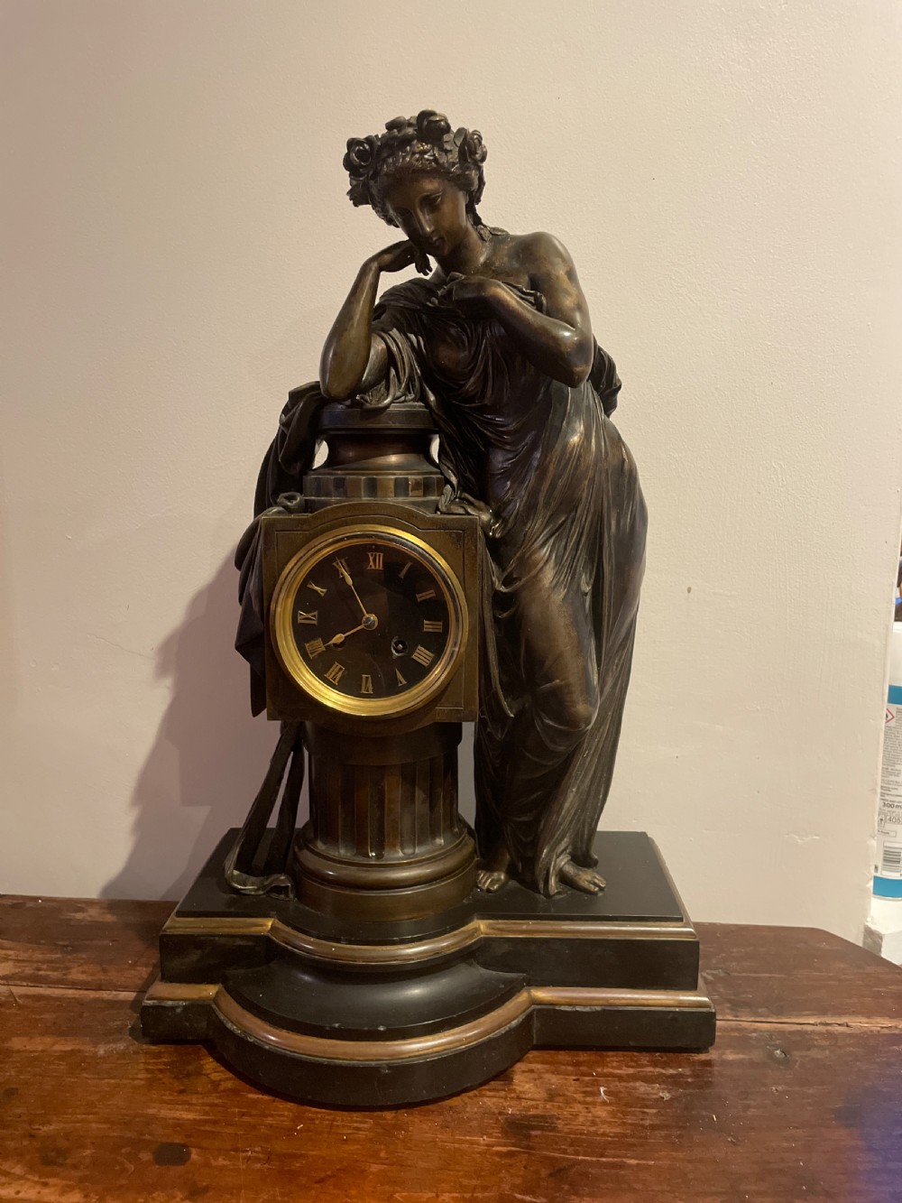 A late 19th-century French dark patinated bronze mantel clock depicting a classical draped female figure leaning upon a column containing a round dial with Roman numerals. View XVII