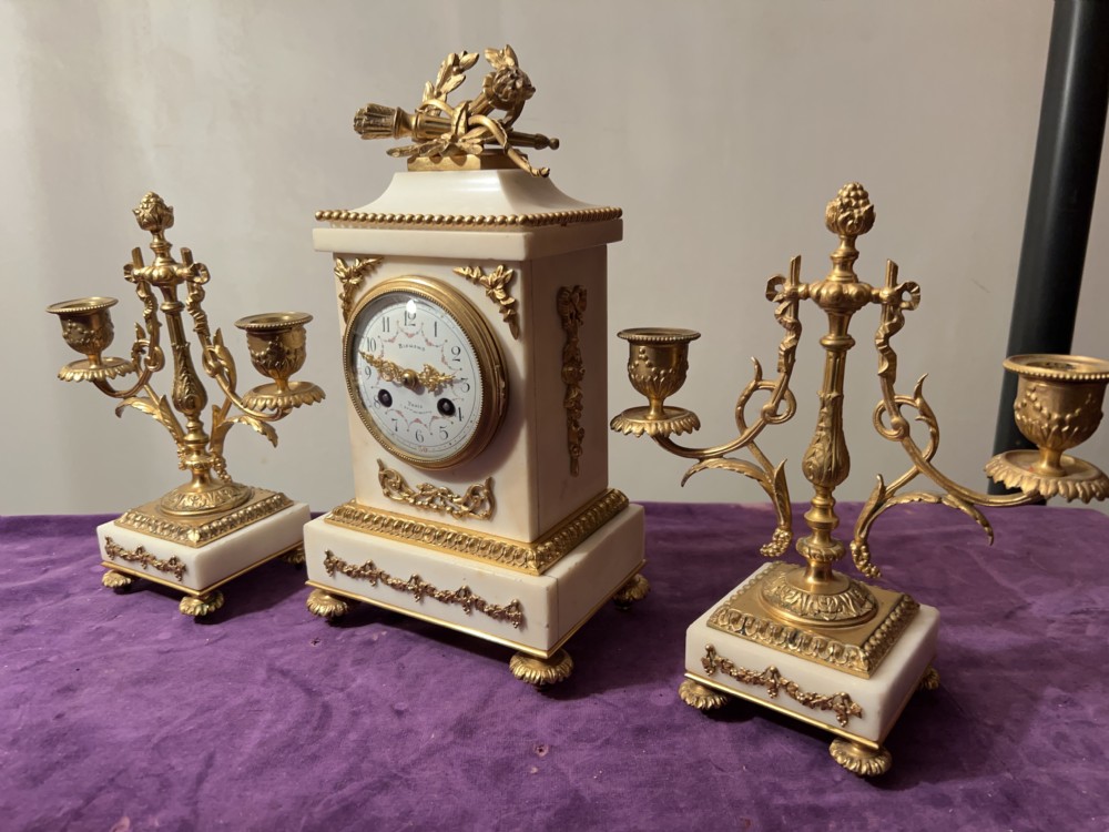 A three-piece French white marble and gilt bronze clock garniture set featuring a central mantel clock topped with a quiver finial alongside two matching candelabra.