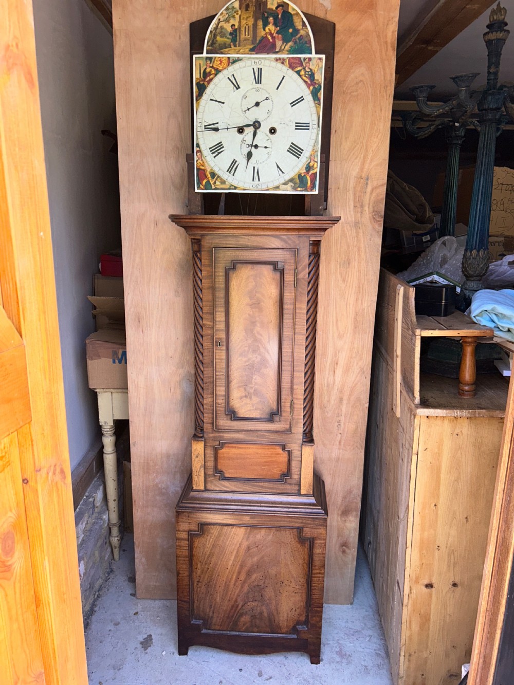 An upright wooden longcase clock featuring a carved swan-neck pediment, a painted dial face, spiral barley twist columns, and a rectangular trunk door. View III
