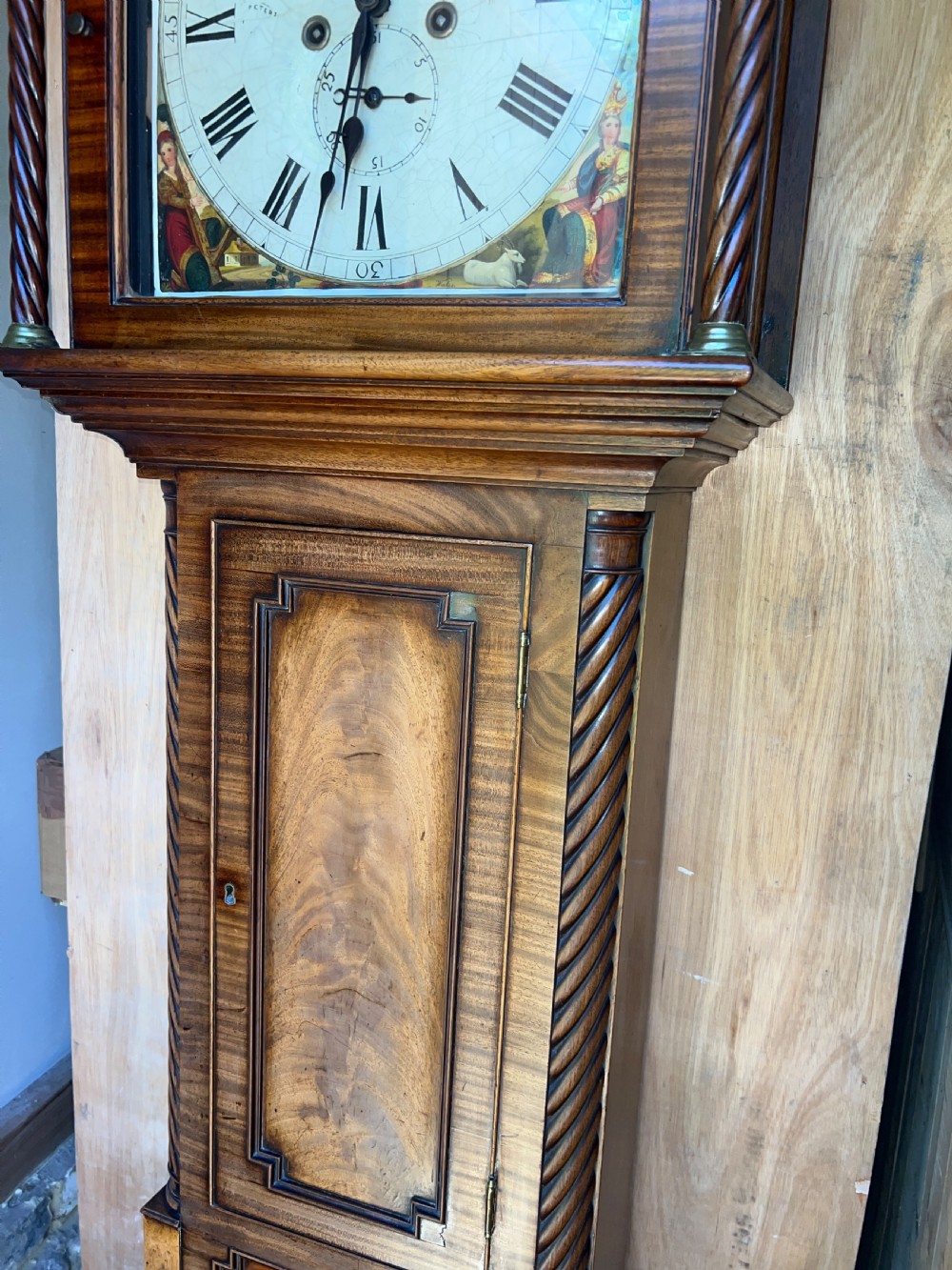 An upright wooden longcase clock featuring a carved swan-neck pediment, a painted dial face, spiral barley twist columns, and a rectangular trunk door. View X