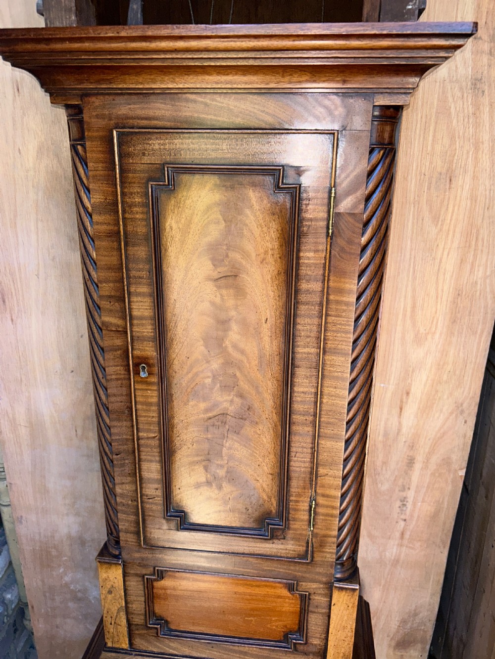 An upright wooden longcase clock featuring a carved swan-neck pediment, a painted dial face, spiral barley twist columns, and a rectangular trunk door. View XIX