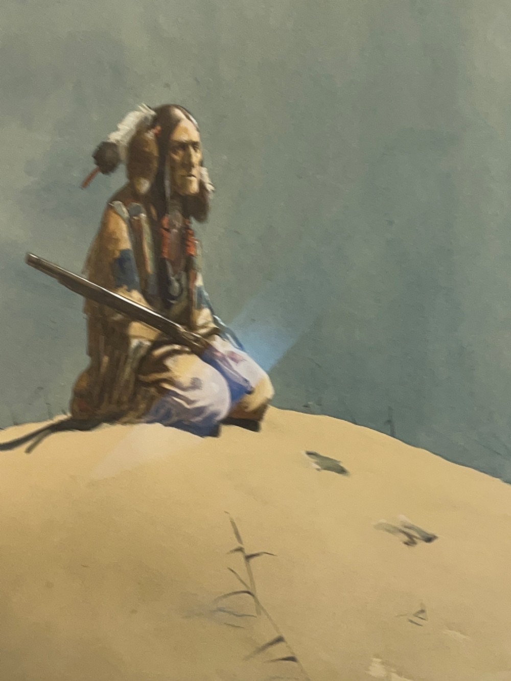 A framed watercolour painting depicting a Native American figure seated on a grassy mound holding a rifle against a muted blue sky. View V