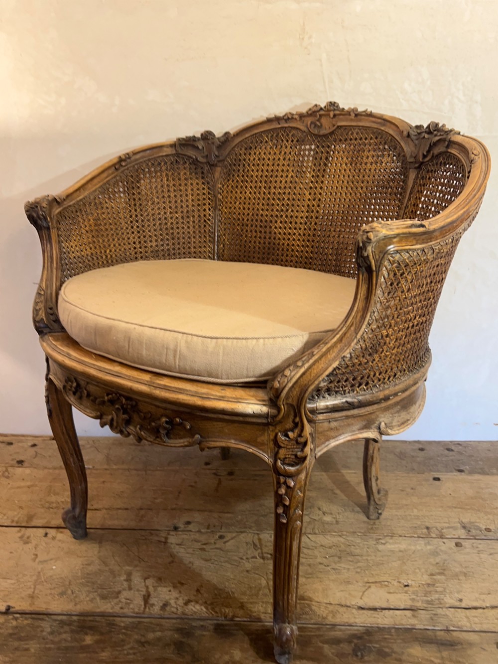 A 19th-century French Louis XV style Corbeille armchair featuring a curved hand-woven cane backrest, carved wooden frame with cabriole legs, and a beige upholstered seat cushion.