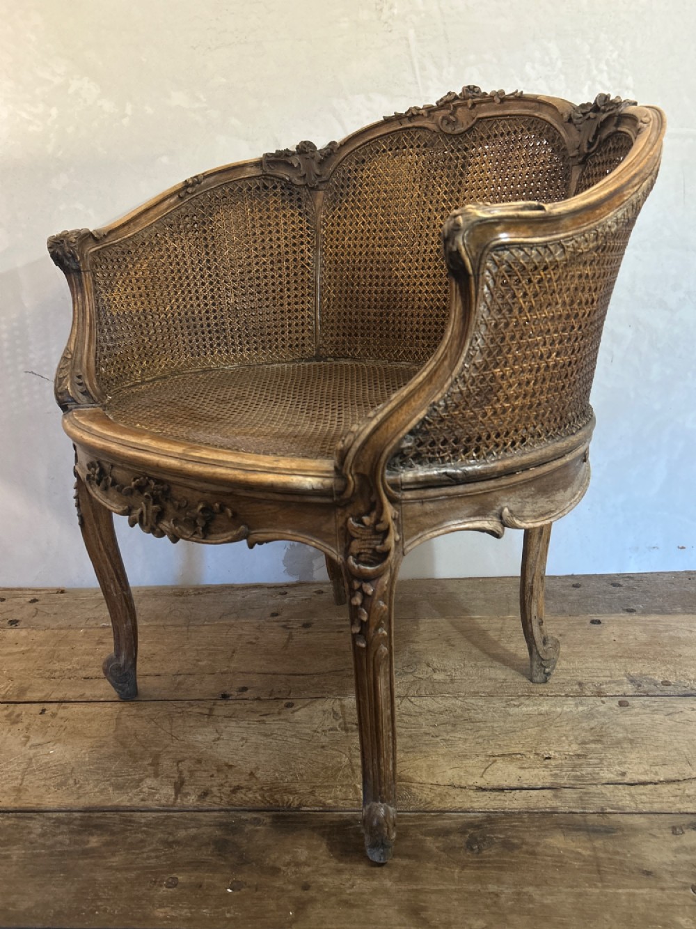 A 19th-century French Louis XV style Corbeille armchair featuring a curved hand-woven cane backrest, carved wooden frame with cabriole legs, and a beige upholstered seat cushion. View II