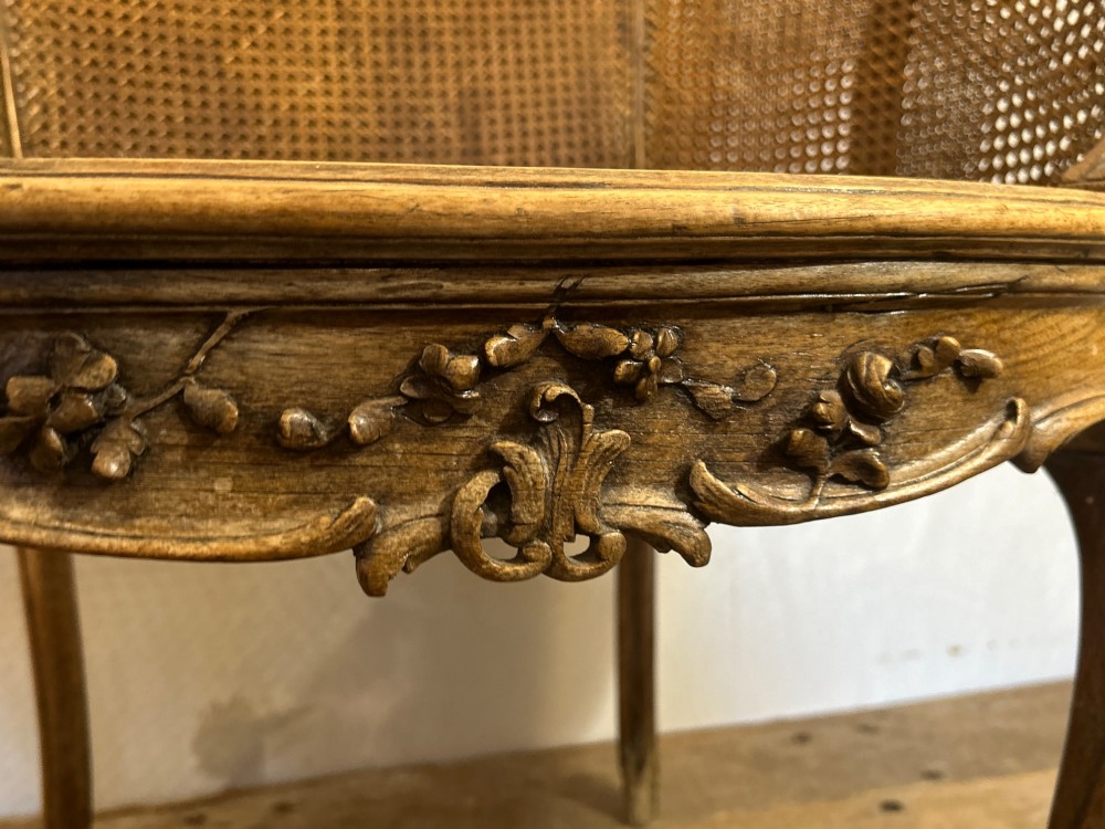 A 19th-century French Louis XV style Corbeille armchair featuring a curved hand-woven cane backrest, carved wooden frame with cabriole legs, and a beige upholstered seat cushion. View VI