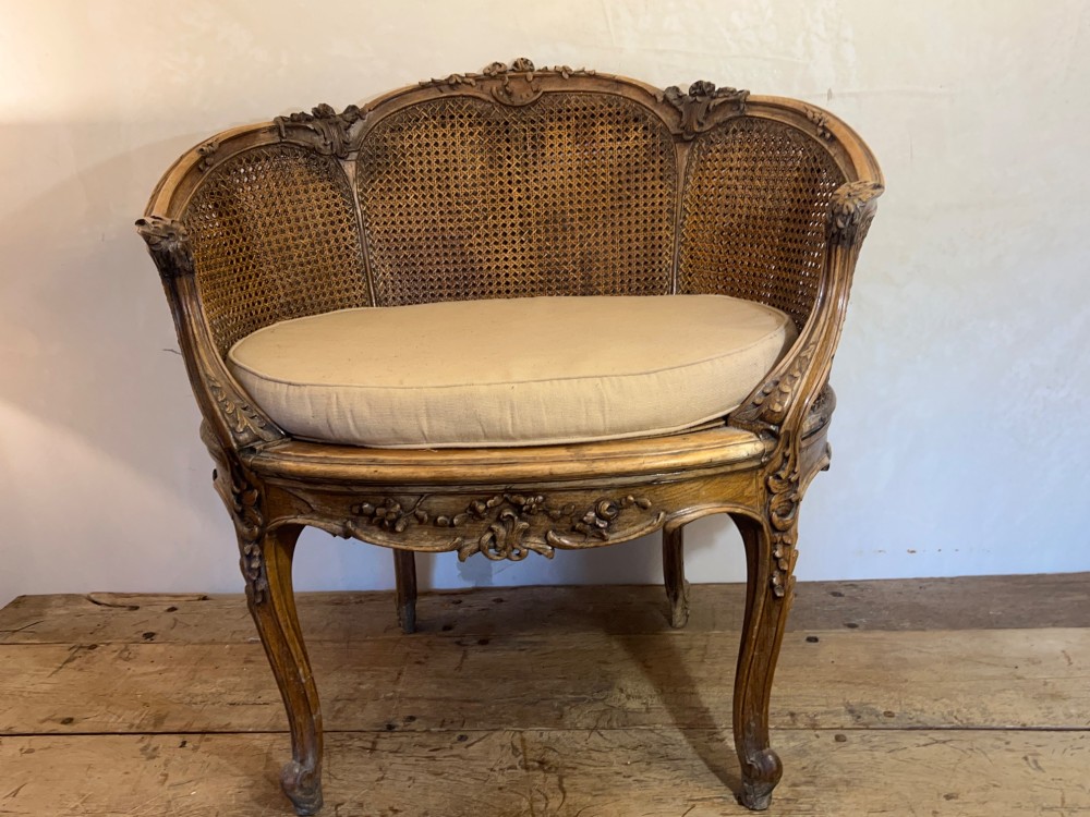 A 19th-century French Louis XV style Corbeille armchair featuring a curved hand-woven cane backrest, carved wooden frame with cabriole legs, and a beige upholstered seat cushion. View X